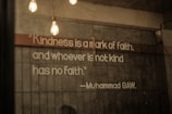 A quote is displayed on a textured concrete wall, illuminated by hanging Edison bulb lights. The text emphasizes the importance of kindness and is attributed to Muhammad SAW.