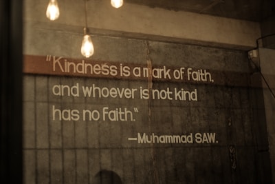 A quote is displayed on a textured concrete wall, illuminated by hanging Edison bulb lights. The text emphasizes the importance of kindness and is attributed to Muhammad SAW.
