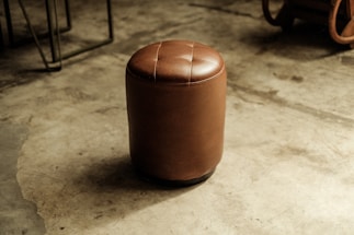 A brown leather cylindrical stool with a button-tufted top situated on a polished concrete floor. The background includes parts of metal and wooden furniture or objects.