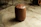 A brown leather cylindrical stool with a button-tufted top situated on a polished concrete floor. The background includes parts of metal and wooden furniture or objects.