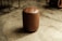 A brown leather cylindrical stool with a button-tufted top situated on a polished concrete floor. The background includes parts of metal and wooden furniture or objects.