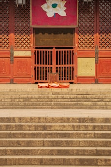 Wooden temple doors with intricate latticework stand atop a series of wide stone steps. A lotus painting adorns the top of the doorway. Three orange cushions are placed in front of the doors, suggesting a space for meditation or prayer.