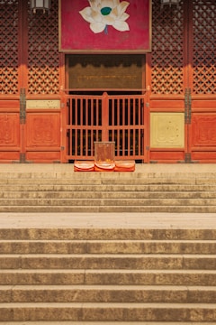 Wooden temple doors with intricate latticework stand atop a series of wide stone steps. A lotus painting adorns the top of the doorway. Three orange cushions are placed in front of the doors, suggesting a space for meditation or prayer.