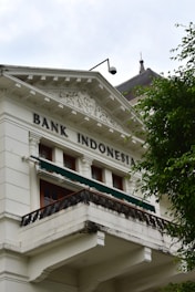 A historic building with classical architecture featuring ornate decorative elements, white facade, and a balcony. The words 'Bank Indonesia' are prominently displayed on the building. A tree with lush green leaves partially obscures the building.