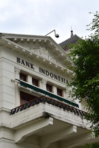 A historic building with classical architecture featuring ornate decorative elements, white facade, and a balcony. The words 'Bank Indonesia' are prominently displayed on the building. A tree with lush green leaves partially obscures the building.