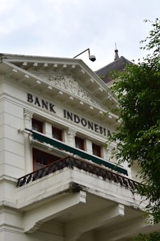 A historic building with classical architecture featuring ornate decorative elements, white facade, and a balcony. The words 'Bank Indonesia' are prominently displayed on the building. A tree with lush green leaves partially obscures the building.