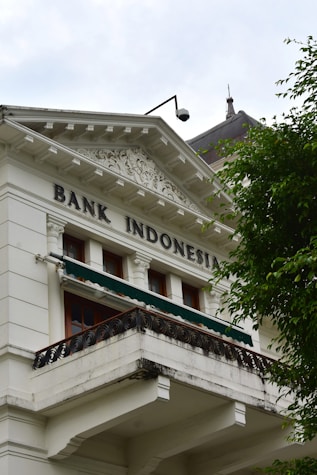 A historic building with classical architecture featuring ornate decorative elements, white facade, and a balcony. The words 'Bank Indonesia' are prominently displayed on the building. A tree with lush green leaves partially obscures the building.
