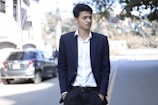 A young man is standing on a street, dressed in a dark suit with a white shirt. He has short, dark hair and his hands are in his pockets. In the background, there is a blurred image of a parked car and some trees overshadowing the street.