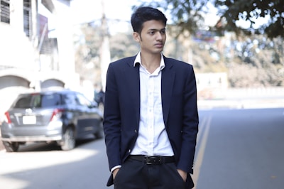 A young man is standing on a street, dressed in a dark suit with a white shirt. He has short, dark hair and his hands are in his pockets. In the background, there is a blurred image of a parked car and some trees overshadowing the street.