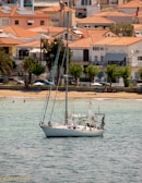 A sailboat is anchored in calm waters near a sandy beach, with a backdrop of Mediterranean-style houses with red-tiled roofs. A few people relax on the beach, some under umbrellas. The sky is clear, suggesting a sunny day.