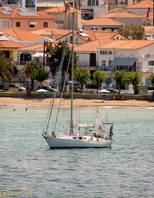 A sailboat is anchored in calm waters near a sandy beach, with a backdrop of Mediterranean-style houses with red-tiled roofs. A few people relax on the beach, some under umbrellas. The sky is clear, suggesting a sunny day.