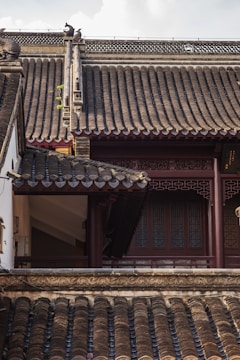 An intricately designed traditional Asian roof features layered, curved tiles with ornate carvings and decorative elements. The structure showcases wooden beams and decorative latticework windows, highlighting a blend of craftsmanship and architectural beauty. The lighting casts soft shadows, enhancing the texture of the roofing tiles.