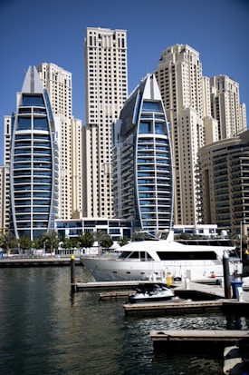 Tall, modern skyscrapers with unique architectural designs are situated near a marina. A luxury yacht is docked on the water, surrounded by smaller boats and docks. The sky is clear and blue.