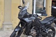 A black motorcycle with a sleek design is parked on a cobblestone surface. It features an angular windshield, detailed engine parts, and a sturdy frame. In the background, a storefront displays a list of services and a small statue, which looks decorative, is placed near the window.