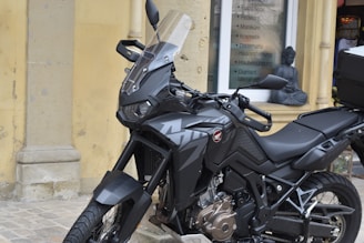 A black motorcycle with a sleek design is parked on a cobblestone surface. It features an angular windshield, detailed engine parts, and a sturdy frame. In the background, a storefront displays a list of services and a small statue, which looks decorative, is placed near the window.