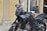 A black motorcycle with a sleek design is parked on a cobblestone surface. It features an angular windshield, detailed engine parts, and a sturdy frame. In the background, a storefront displays a list of services and a small statue, which looks decorative, is placed near the window.