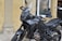 A black motorcycle with a sleek design is parked on a cobblestone surface. It features an angular windshield, detailed engine parts, and a sturdy frame. In the background, a storefront displays a list of services and a small statue, which looks decorative, is placed near the window.