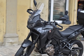 A black motorcycle with a sleek design is parked on a cobblestone surface. It features an angular windshield, detailed engine parts, and a sturdy frame. In the background, a storefront displays a list of services and a small statue, which looks decorative, is placed near the window.