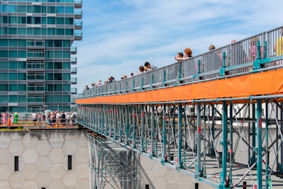 A group of people are walking on a temporary bridge with orange barriers, supported by metal scaffolding. The bridge is in an urban setting, near a modern, glass-windowed building. The sky is blue, and the scene appears crowded yet organized with individuals looking over the railing and some taking photographs.