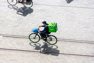 A cyclist, carrying a green delivery bag, is riding a bicycle on a paved street. The bicycle features green rims and a blue front basket or bag. The cyclist is dressed in dark clothing and a cap. There is another cyclist partially visible towards the top of the image. The shadows of both cyclists are cast on the ground.