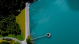 Aerial view of a large body of turquoise water adjacent to a forest on the left. A straight line of a road separates the green forest from a well-maintained grassy area, leading to a round structure on the water resembling a valve or spillway.