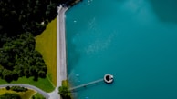 Aerial view of a large body of turquoise water adjacent to a forest on the left. A straight line of a road separates the green forest from a well-maintained grassy area, leading to a round structure on the water resembling a valve or spillway.
