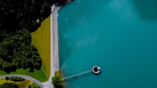 Aerial view of a large body of turquoise water adjacent to a forest on the left. A straight line of a road separates the green forest from a well-maintained grassy area, leading to a round structure on the water resembling a valve or spillway.