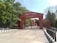 A red entrance gate with ornate details provides access to a significant building complex. Trees with lush greenery surround the area, and the sky is clear and bright. Metal gates are open, and there are some signs and an empty chair visible.