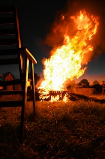 A bright and large fire burns intensely in an outdoor setting, surrounded by grass. A wooden chair is positioned prominently in the foreground to the left, creating silhouettes against the fire's glow. In the background, there are other elements such as hay bales and trees, suggesting a rural or campsite location.