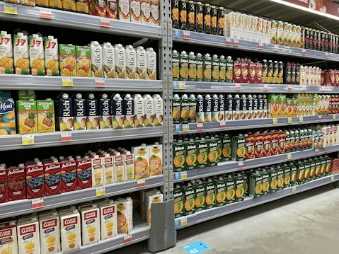 Shelves stocked with various brands and flavors of packaged fruit juices. The cartons are neatly arranged and display a variety of fruit illustrations, indicating different juice flavors. The shelves are labeled with price tags, and the atmosphere resembles a well-organized grocery store aisle.