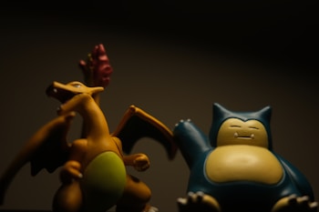 Two collectible figurines are positioned side by side in a dimly lit environment, casting subtle shadows. The first is a dragon with an orange body, wings extended, and a greenish belly. The second figure resembles a large, blue and cream-colored creature with a sleepy expression. Both figures are depicted with detailed features.