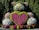 A large topiary sculpture of a teddy bear is beautifully adorned with vibrant flowers and greenery. The bear features a heart made of bright pink and red flowers prominently on its chest, surrounded by green hedges. The sculpture is set against a backdrop of lush trees, enhancing its vivid appearance.