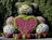 A large topiary sculpture of a teddy bear is beautifully adorned with vibrant flowers and greenery. The bear features a heart made of bright pink and red flowers prominently on its chest, surrounded by green hedges. The sculpture is set against a backdrop of lush trees, enhancing its vivid appearance.