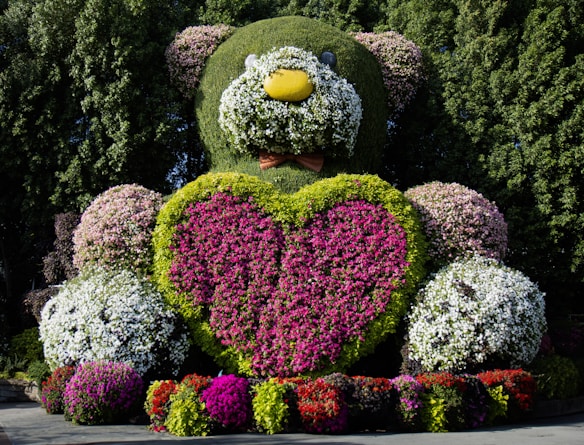 A large topiary sculpture of a teddy bear is beautifully adorned with vibrant flowers and greenery. The bear features a heart made of bright pink and red flowers prominently on its chest, surrounded by green hedges. The sculpture is set against a backdrop of lush trees, enhancing its vivid appearance.
