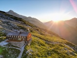 A secluded mountain cabin sits on a grassy hillside at sunset, with rugged mountains and rocks surrounding it. The warm sunlight bathes the landscape, creating a serene and peaceful atmosphere.