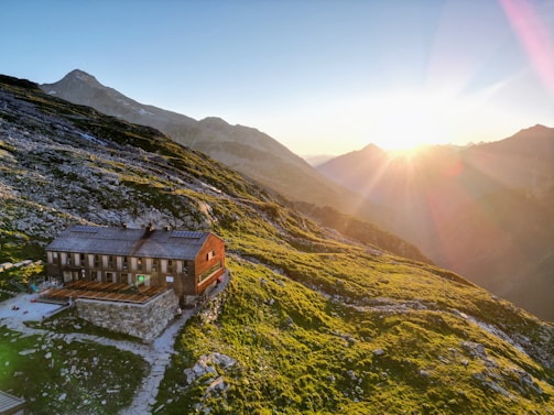 A secluded mountain cabin sits on a grassy hillside at sunset, with rugged mountains and rocks surrounding it. The warm sunlight bathes the landscape, creating a serene and peaceful atmosphere.