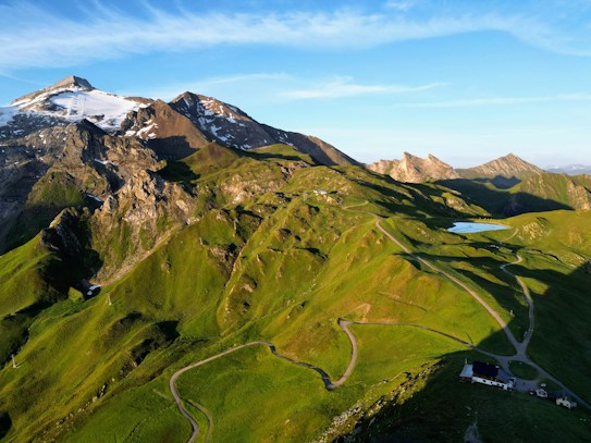 A breathtaking mountainous landscape featuring lush green hills and winding paths. Snow-capped peaks and rocky surfaces tower in the distance under a clear blue sky. The sunlight casts dramatic shadows, enhancing the varied terrain. A small building is nestled at the foot of the hills alongside a serene blue lake.