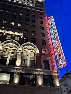 A multistory brick building with ornate architectural details like arched windows and decorative columns, illuminated by warm lights. A large neon sign displaying the word 'Proper Hotel' is affixed to the facade, glowing against the deep blue evening sky.