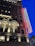 A multistory brick building with ornate architectural details like arched windows and decorative columns, illuminated by warm lights. A large neon sign displaying the word 'Proper Hotel' is affixed to the facade, glowing against the deep blue evening sky.