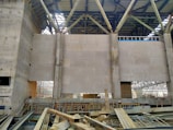 The interior of an unfinished industrial building with concrete walls and metal beams. Large beige panels form part of the walls, while exposed structures reveal scaffolding and construction materials scattered on the ground. The roof is partially completed with metal trusses.