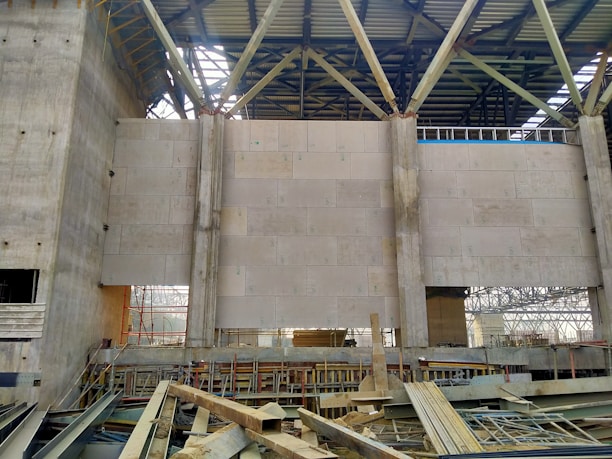 The interior of an unfinished industrial building with concrete walls and metal beams. Large beige panels form part of the walls, while exposed structures reveal scaffolding and construction materials scattered on the ground. The roof is partially completed with metal trusses.