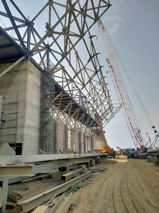 A construction site with a large, unfinished steel structure supported by concrete pillars. A crane is lifting materials to the upper part of the structure. Scaffolding is set up on the left side of the site. Piles of steel beams and other materials lie on the ground in front of the structure. The ground appears sandy, and the sky is clear.