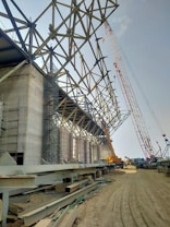 A construction site with a large, unfinished steel structure supported by concrete pillars. A crane is lifting materials to the upper part of the structure. Scaffolding is set up on the left side of the site. Piles of steel beams and other materials lie on the ground in front of the structure. The ground appears sandy, and the sky is clear.
