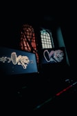 A dimly lit room features two illuminated computer screens displaying stylized graphics of an animal with elements of graffiti art and Japanese text. A window with a grid design is partially obscured by a translucent curtain, casting subtle patterns of light and shadow.