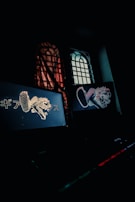 A dimly lit room features two illuminated computer screens displaying stylized graphics of an animal with elements of graffiti art and Japanese text. A window with a grid design is partially obscured by a translucent curtain, casting subtle patterns of light and shadow.