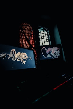 A dimly lit room features two illuminated computer screens displaying stylized graphics of an animal with elements of graffiti art and Japanese text. A window with a grid design is partially obscured by a translucent curtain, casting subtle patterns of light and shadow.