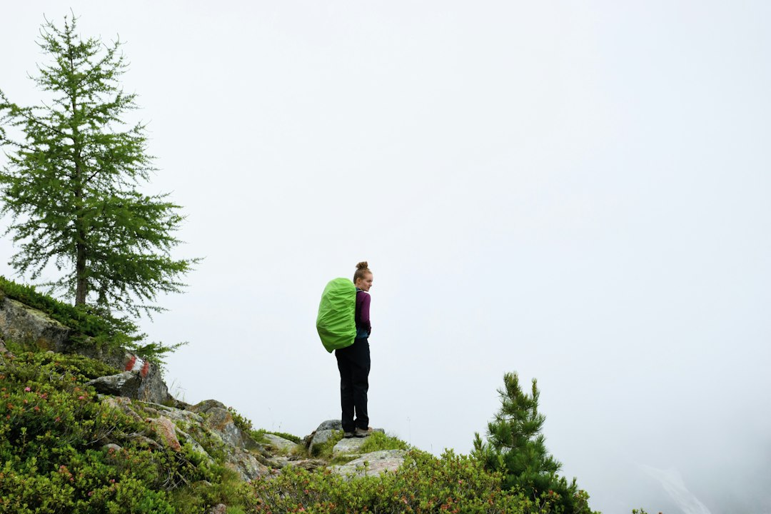 a person with a green backpack standing on a hill, A girl standing with a backpack in the Austrian Alps looking at the m