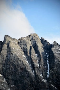Sharp peaks of rocky mountains with patches of snow are seen against a blue sky with some clouds.