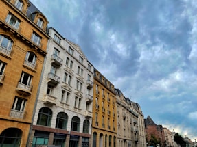 A row of European-style buildings, featuring ornate architectural details, stretches along a street beneath a cloudy sky. The buildings have multiple stories with large windows and balconies, displaying a mix of beige, white, and light yellow facades.