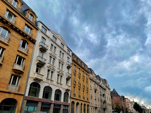 A row of European-style buildings, featuring ornate architectural details, stretches along a street beneath a cloudy sky. The buildings have multiple stories with large windows and balconies, displaying a mix of beige, white, and light yellow facades.